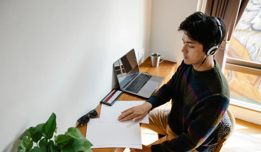 A man working at his laptop with a plant, coffee mug, and sunglasses on his desk. It's a bright sunny day.