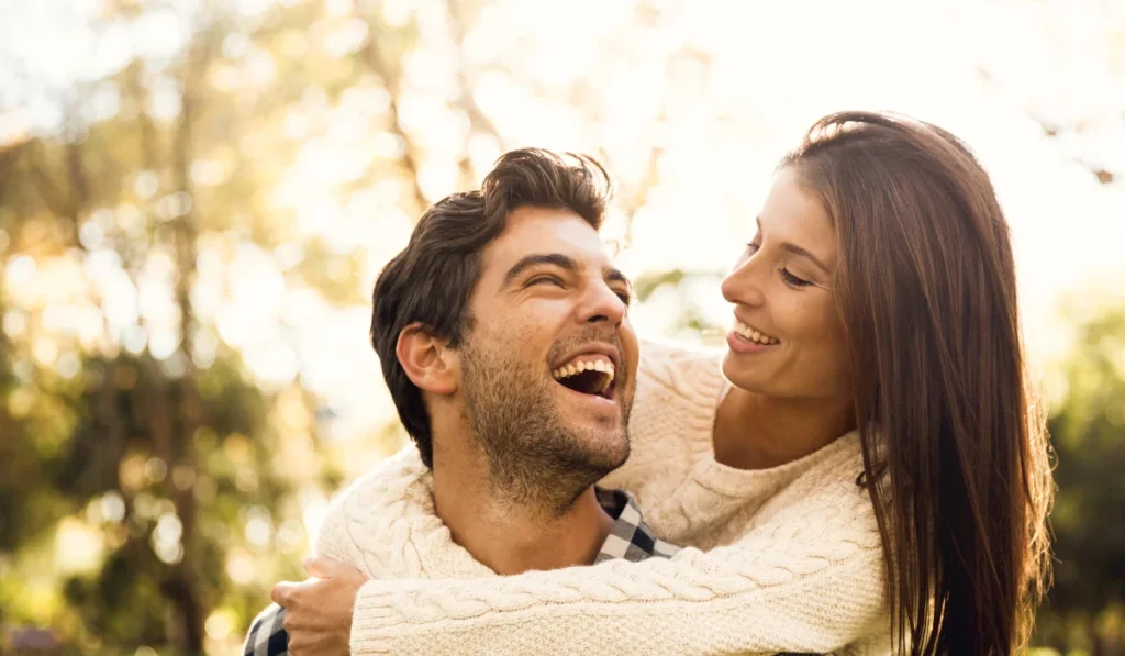 A happy couple smiling at each other. It's a sunny day and she's wrapped her arms around him from behind.