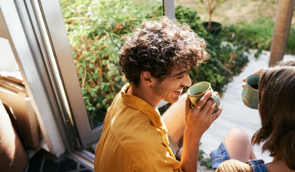 A couple sitting on the porch on a bright sunny day drinking coffee together and smiling as they chat.