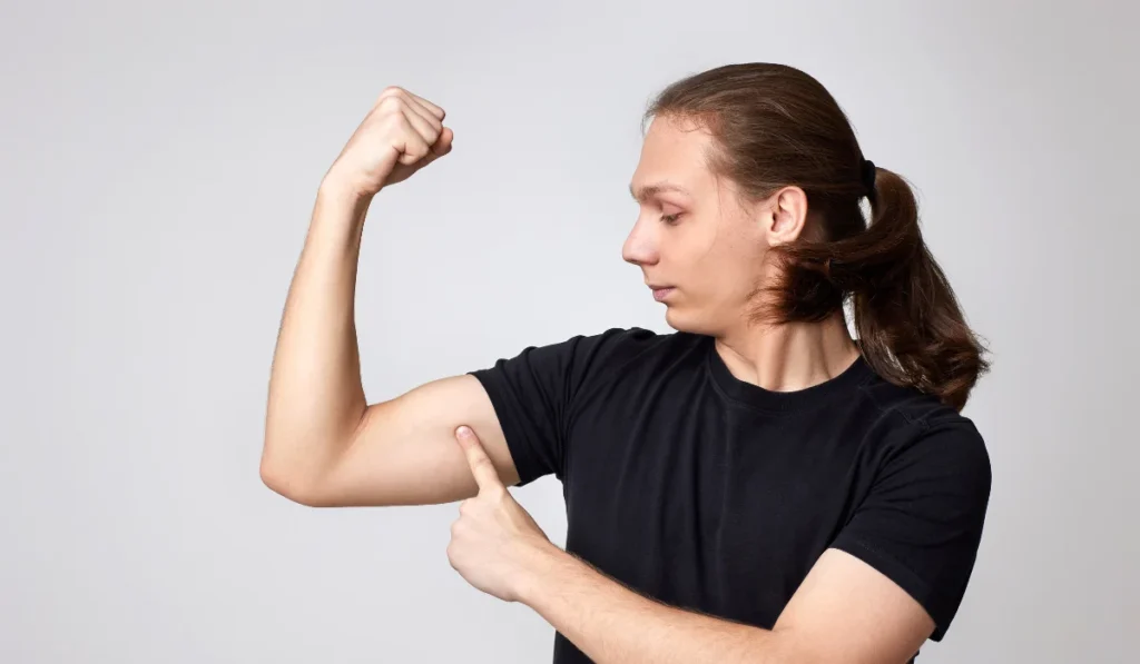 A genderfluid person flexing their bicep. They have long brown hair in a ponytail and are wearing a black t-shirt.