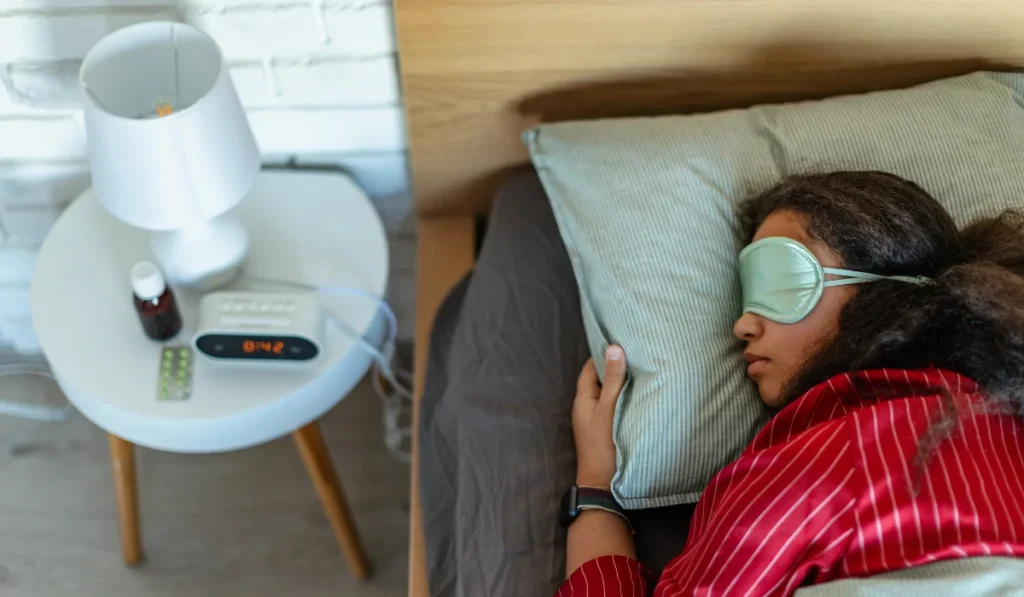 A young woman asleep with a sleeping mask on. There are pills on her nightstand.