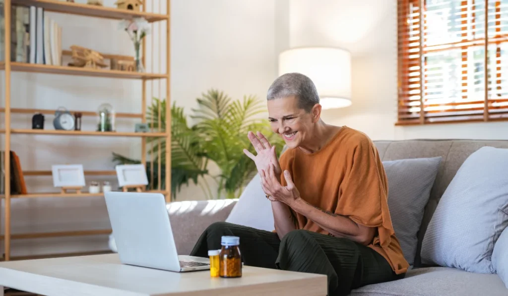 An older woman with a shaved head smiling and waving at her laptop screen. There are pills on her coffee table next to the laptop.