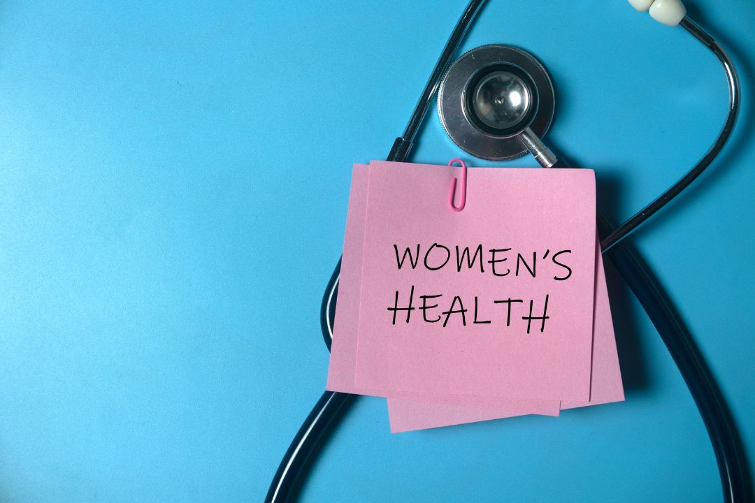 A stethoscope and pink post-it note with "women's health" written on it on a blue background.